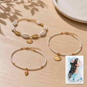 May include: Three bracelets with gold-toned metal shell charms. One bracelet features white cowrie shells and gold-toned shells. Two bracelets consist of small white beads with gold-toned accents. A card shows a watercolor illustration with text.