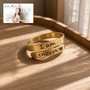 K&ouml;nnte beinhalten: Ein goldfarbener Wickelring mit der Gravur I am enough und einem kleinen Herzsymbol. Der Ring hat eine glatte, polierte Oberfl&auml;che und ein offenes Design. Ein kleines Bild zeigt ein Paar mit dem Text forever with you Olivia & Adam.
