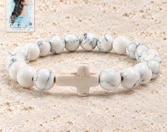 Könnte beinhalten: Ein elastisches Armband mit runden weißen Perlen mit grauer Maserung und einer zentralen, horizontalen kreuzförmigen Perle. Enthalten ist eine kleine rechteckige Karte mit einer Illustration einer Person und dem Text "forever with you".