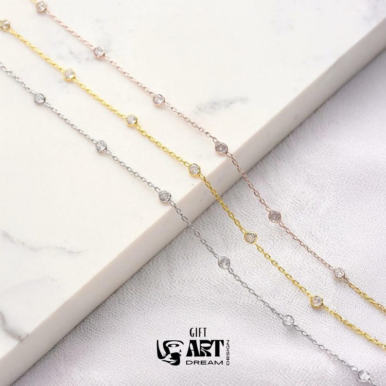 May include: Four delicate necklaces in silver, gold, and rose gold tones, each featuring small, clear gemstones evenly spaced along the chain. The necklaces are displayed on a white surface, with the Gift Art Dream Design logo visible.