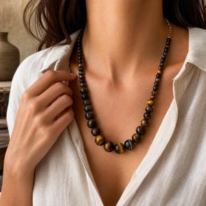 May include: A beaded necklace featuring round, polished tiger's eye stones in shades of golden brown, dark brown, and deep blue. The beads are graduated in size, with the largest stones centered at the bottom and smaller beads extending upward.