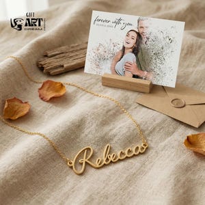 May include: A gold-colored necklace with the name "Rebecca" in cursive script. A greeting card with a photo of a couple and the text "forever with you" is displayed on a wooden stand. An envelope and dried petals are also present.