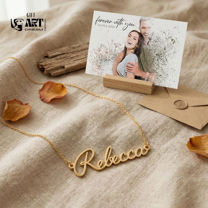 May include: A gold-colored necklace with the name "Rebecca" in cursive script. A greeting card with a photo of a couple and the text "forever with you" is displayed on a wooden stand. An envelope and dried petals are also present.