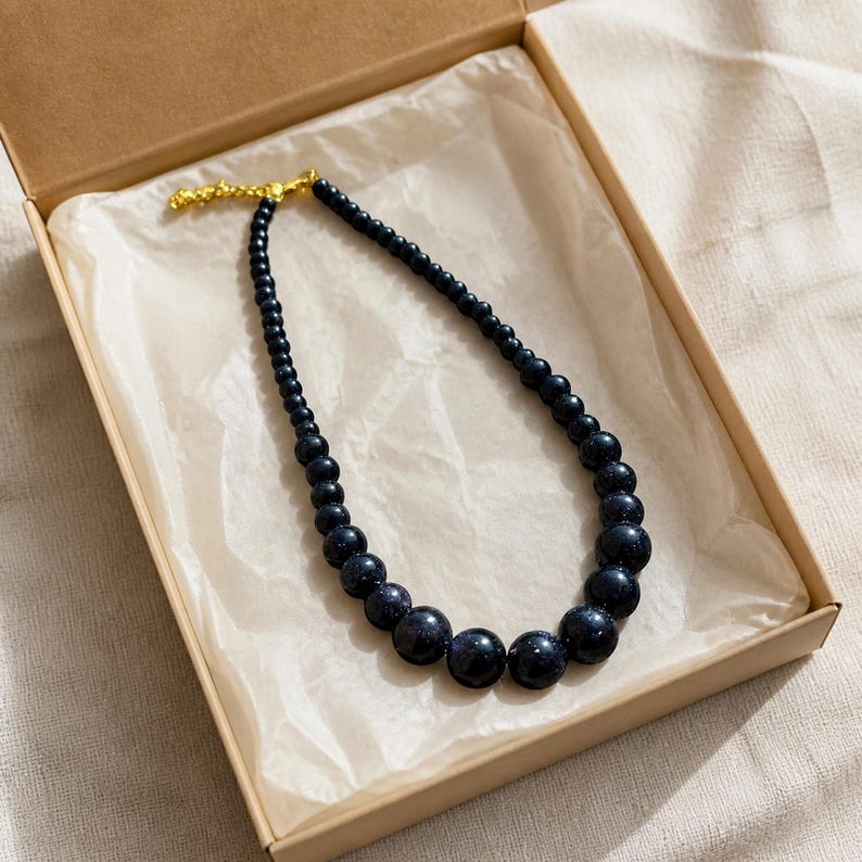 May include: A necklace featuring dark blue, glittery, spherical beads that gradually increase in size toward the center. The strand is secured with a gold-toned metal clasp and is presented inside a light brown cardboard gift box.
