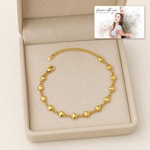 May include: A gold-toned chain bracelet featuring a series of small, polished heart-shaped charms linked together. The bracelet includes an adjustable extender chain with a lobster claw clasp, presented inside a beige jewelry gift box.