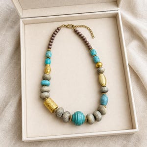 May include: A beaded necklace featuring a mix of turquoise-colored stone beads, matte grey round beads, and gold-toned metal accents. The design includes a central ribbed turquoise bead, cylindrical gold-toned beads, and a lobster claw clasp.