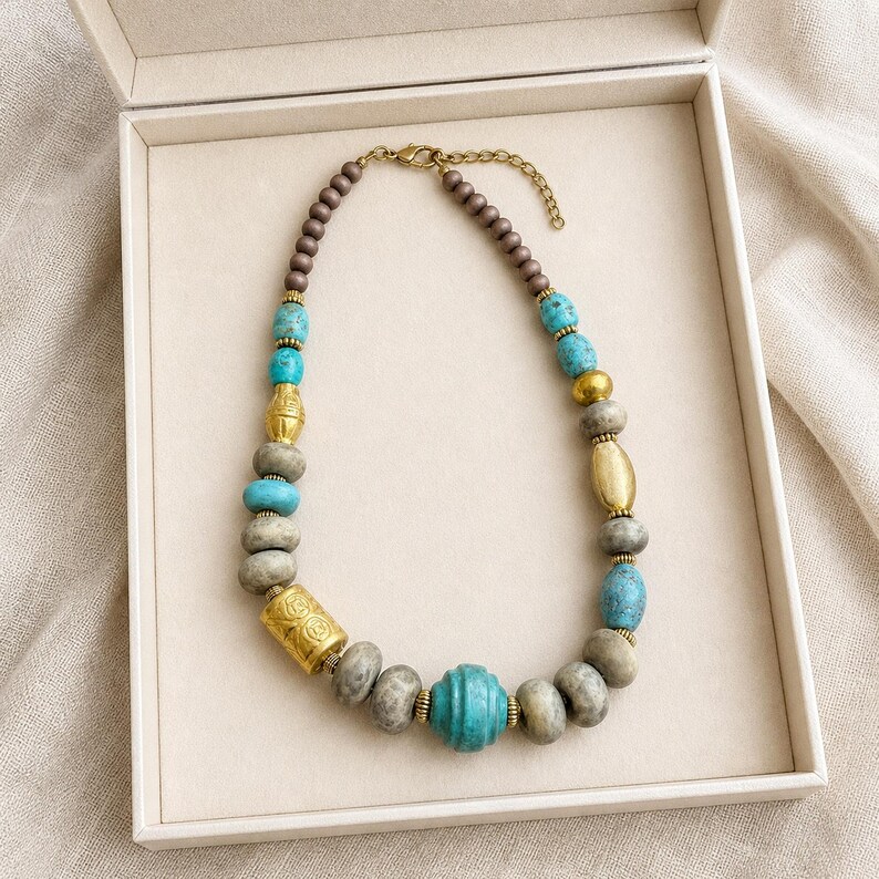 May include: A beaded necklace featuring a mix of turquoise-colored stone beads, matte grey round beads, and gold-toned metal accents. The design includes a central ribbed turquoise bead, cylindrical gold-toned beads, and a lobster claw clasp.