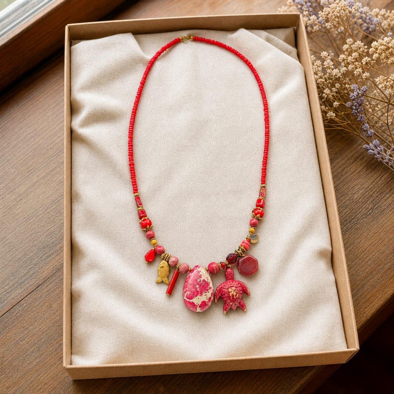 May include: A beaded necklace featuring a vibrant red strand accented with various charms, including a gold-toned fish, a pink and white marbled teardrop stone, and a red turtle figure, all arranged on a light beige fabric lining inside a box.