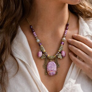Puede incluir: Un collar con un gran colgante ovalado de color morado moteado, espaciadores de metal en tono dorado antiguo y una hilera de cuentas moradas, lavanda y transl&uacute;cidas. El dise&ntilde;o incluye cadenas met&aacute;licas colgantes y detalles de filigrana.
