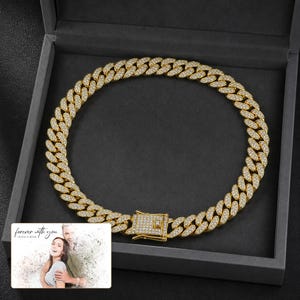 May include: A gold-toned cuban link chain necklace featuring a pav&eacute; setting with numerous small clear stones embedded across the surface of each link and a rectangular box clasp closure, presented inside a dark gray jewelry display box.