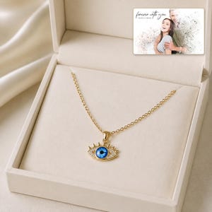 Gold-tone necklace with a blue eye-shaped pendant, presentation in an open jewelry box. A personalized greeting-card example appears in the corner.