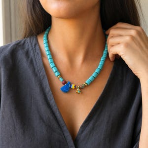 May include: A beaded necklace featuring cylindrical turquoise-colored beads, accented with a small blue tassel, a multi-colored striped bead, a gold-toned charm, and a small patterned bead, all strung together on a cord worn around the neck.