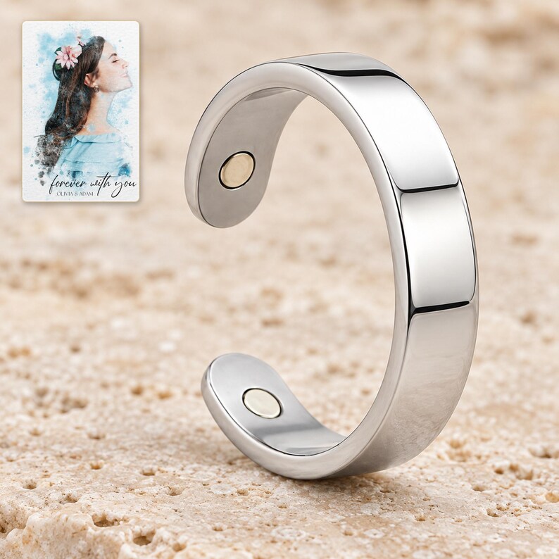 Close upright view of a polished silver-tone open ring on pale travertine, showing its smooth band and two small inner-tip inserts. A personalized digital greeting card is shown alongside.