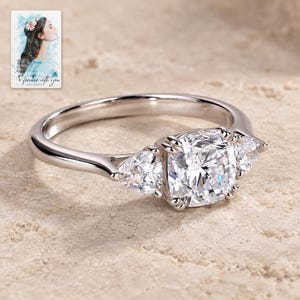 May include: A silver-toned ring featuring a central cushion-cut stone flanked by two smaller pear-shaped stones. The band is smooth and polished, holding the three clear, faceted stones in a prong setting. The design is clean and symmetrical.