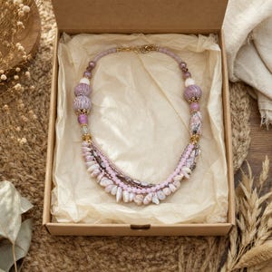 May include: A multi-strand necklace featuring a variety of beads in shades of lavender, mauve, and soft pink. The design includes textured round beads, smooth spherical beads, and small irregular shell-like components, all connected by gold-tone metal accents.