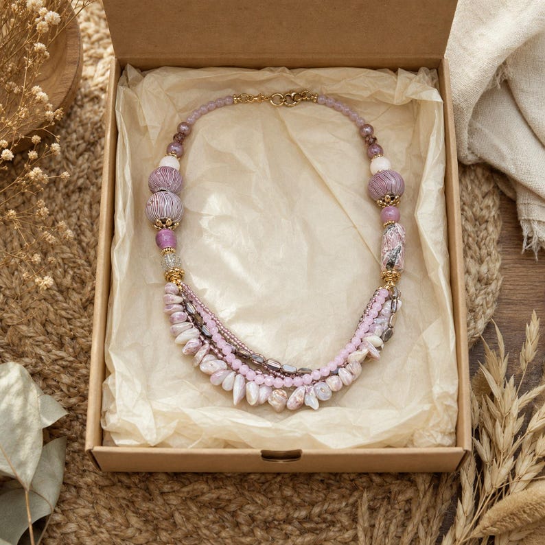 May include: A multi-strand necklace featuring a variety of beads in shades of lavender, mauve, and soft pink. The design includes textured round beads, smooth spherical beads, and small irregular shell-like components, all connected by gold-tone metal accents.