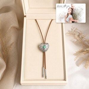 Brown cord bolo necklace with a silver-tone heart slide and turquoise-color center, presentation in an open jewelry box. A personalized greeting-card example appears in the corner.