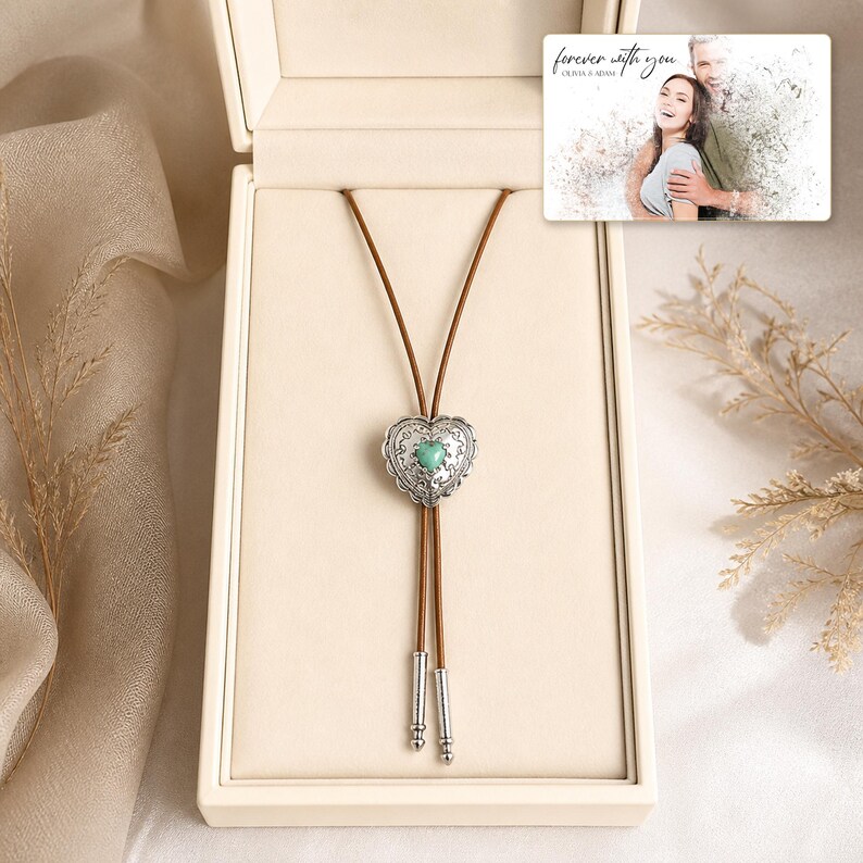 Brown cord bolo necklace with a silver-tone heart slide and turquoise-color center, presentation in an open jewelry box. A personalized greeting-card example appears in the corner.