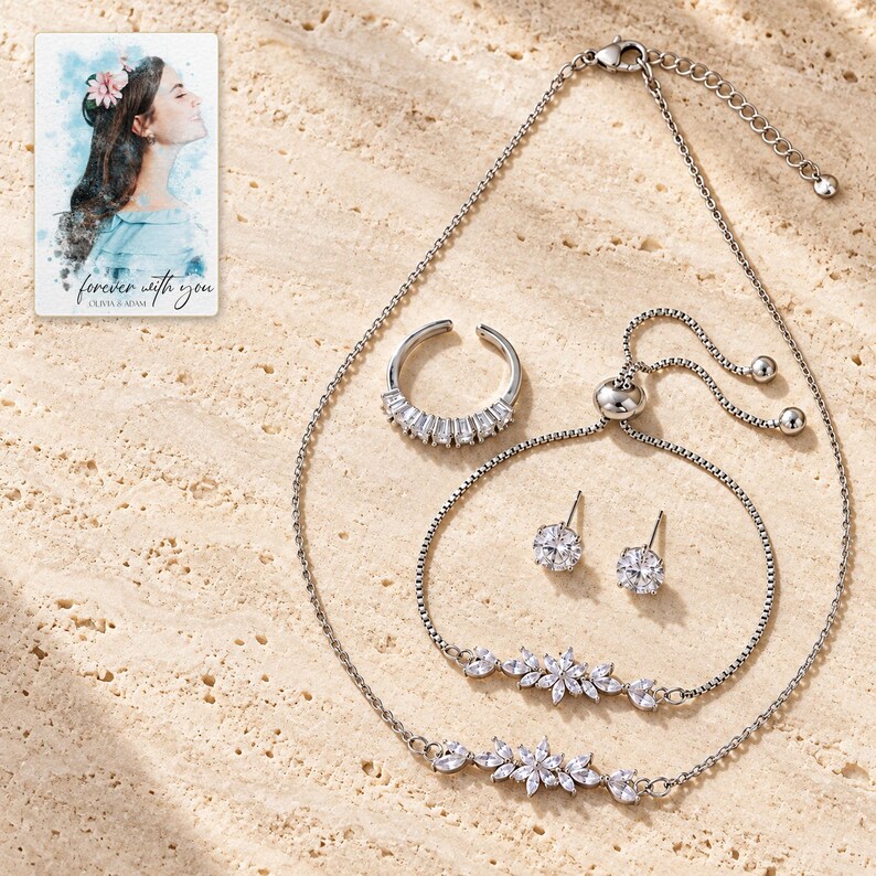 Silver-tone floral jewelry set on ivory stone style with leaf necklace, matching bolo bracelet, open baguette ring and two clear round stud earrings. A personalized digital greeting card is shown alongside.