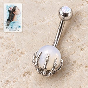 Complete silver-tone curved navel barbell with a white ball held in a skeleton-hand cradle on pale stone style. A personalized digital greeting card is shown alongside.