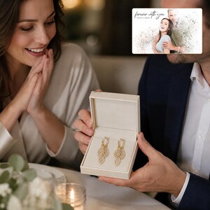 May include: A pair of gold hoop earrings featuring dangling leaf-shaped charms encrusted with small clear stones, presented in a cream-colored rectangular jewelry box. The earrings are displayed against a soft, light-colored interior lining.