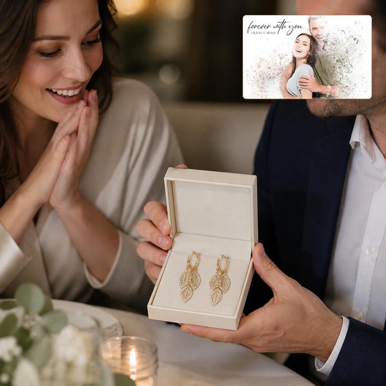 May include: A pair of gold hoop earrings featuring dangling leaf-shaped charms encrusted with small clear stones, presented in a cream-colored rectangular jewelry box. The earrings are displayed against a soft, light-colored interior lining.