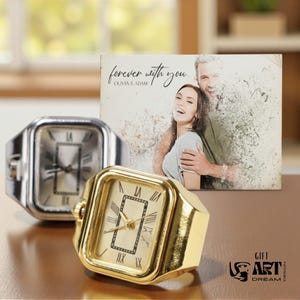 May include: Two square-faced ring watches, one silver and one gold, with Roman numeral hour markers. A photo with the text "forever with you" and a couple is in the background. The watches are on a brown surface.