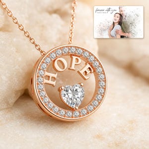 Puede incluir: Un collar de oro rosa con un colgante circular que presenta la palabra HOPE en letras may&uacute;sculas y una piedra transparente en forma de coraz&oacute;n en el centro. El marco circular est&aacute; incrustado con peque&ntilde;as piedras. Una tarjeta muestra una foto con el texto forever with you.