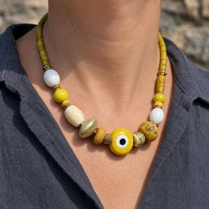 Mustard yellow white and antique gold chunky necklace with central yellow evil eye bead worn with an ivory linen top in warm natural window light