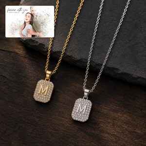 May include: Two rectangular pendant necklaces, one gold-toned and one silver-toned, each featuring a central letter M surrounded by small clear stones. An inset image shows a couple with the text forever with you OLIVIA & ADAM.