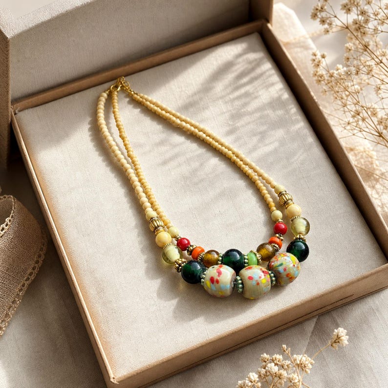 May include: A double-strand necklace featuring small cream-coloured beads, accented with larger round beads in shades of dark green, red, orange, and light green, along with three prominent speckled beads in cream, red, and blue, separated by gold-toned spacers.