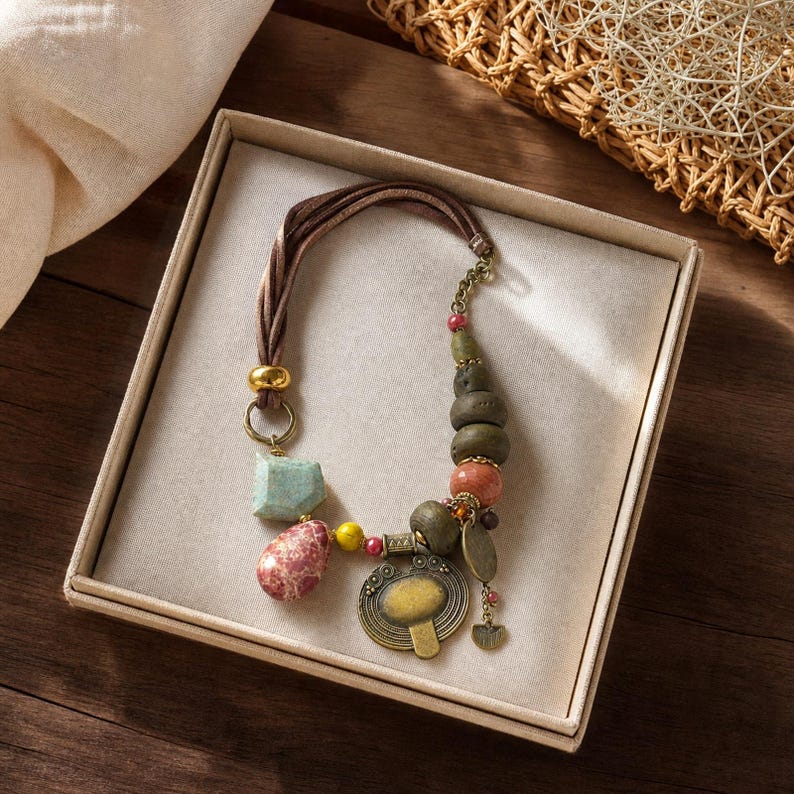May include: A necklace featuring a brown leather cord, a large brass pendant, and various beads in shades of green, pink, and yellow. The piece includes a smooth turquoise-colored stone, a speckled pink stone, and several round, textured olive-green beads.