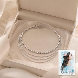May include: A multi-strand silver-toned necklace featuring one row of clear round-cut rhinestones and three delicate beaded chains. The necklace is presented in a cream-coloured box alongside a card with a watercolour illustration and the text "forever with you".