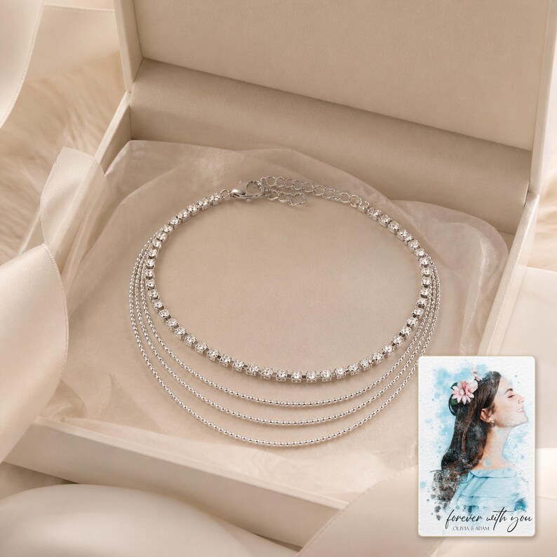 May include: A multi-strand silver-toned necklace featuring one row of clear round-cut rhinestones and three delicate beaded chains. The necklace is presented in a cream-coloured box alongside a card with a watercolour illustration and the text "forever with you".