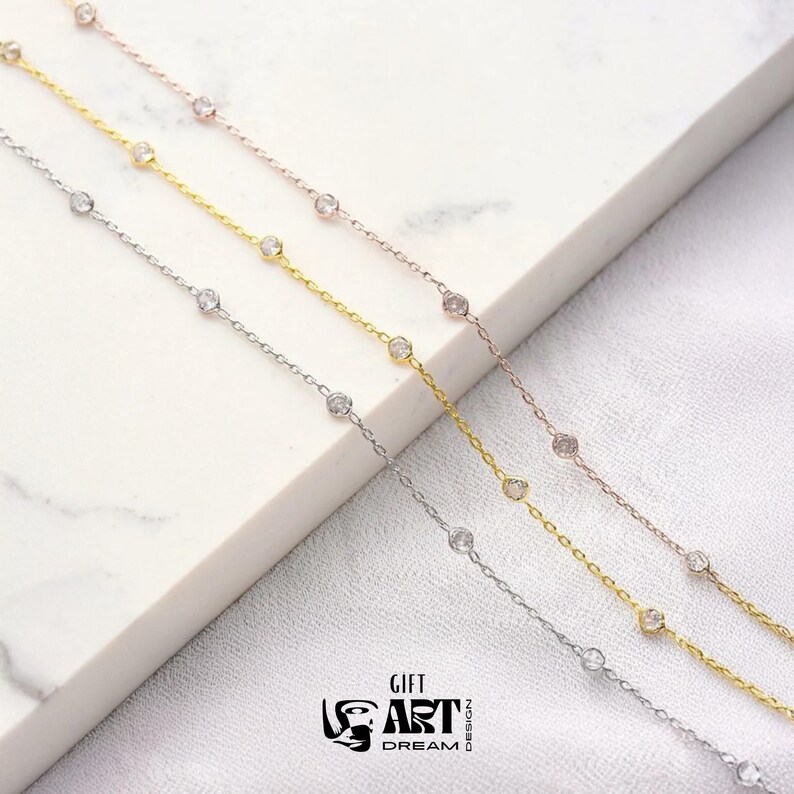 May include: Four delicate necklaces in silver, gold, and rose gold, each featuring small, clear gemstones evenly spaced along the chain. The necklaces are displayed on a white surface, with the logo "GIFT ART DREAM DESIGN" visible.