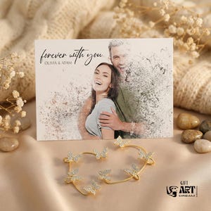 May include: A gold-coloured ear cuff featuring butterfly designs and a card with the text "forever with you" and a couple's photo. The ear cuff has small, sparkling stones on the butterflies. The card is on a soft, neutral-toned background.