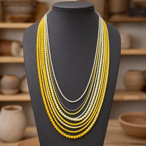 May include: A multi-strand necklace featuring numerous layers of small, round beads in varying shades of yellow, ranging from pale lemon to deep golden yellow, interspersed with thin, metallic silver-toned chains on a dark display bust.
