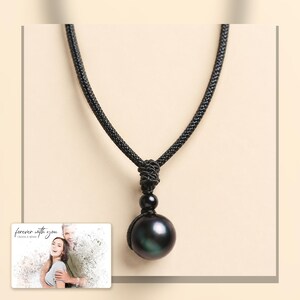 Black cord necklace with a dark round pendant, light-bordered product view. A personalized greeting-card example appears in the corner.