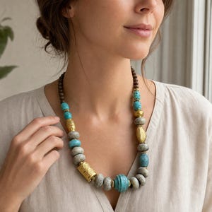May include: A beaded necklace featuring a mix of dark brown wooden beads, round turquoise-colored beads, smooth grey stone-like beads, and textured gold-toned metal cylindrical and spherical spacers arranged in a repeating pattern.