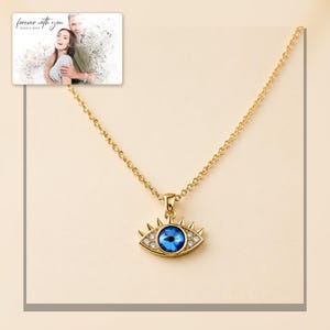Gold-tone necklace with a blue eye-shaped pendant, framed product view. A personalized greeting-card example appears in the corner.