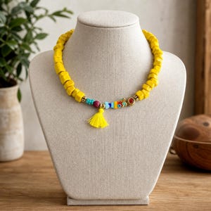 May include: A necklace featuring a strand of bright yellow, irregularly shaped beads. The center includes a small, colorful fish-shaped charm, a red bead, a turquoise bead, and a vibrant yellow tassel hanging from the bottom of the necklace.