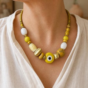 Puede incluir: Un collar de cuentas con un amuleto central de ojo turco amarillo, intercalado con varias cuentas amarillas, blancas y doradas met&aacute;licas. El collar incluye cuentas redondas lisas, cuentas cil&iacute;ndricas texturizadas y peque&ntilde;os espaciadores dorados.