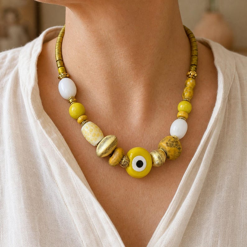 Puede incluir: Un collar de cuentas con un amuleto central de ojo turco amarillo, intercalado con varias cuentas amarillas, blancas y doradas met&aacute;licas. El collar incluye cuentas redondas lisas, cuentas cil&iacute;ndricas texturizadas y peque&ntilde;os espaciadores dorados.