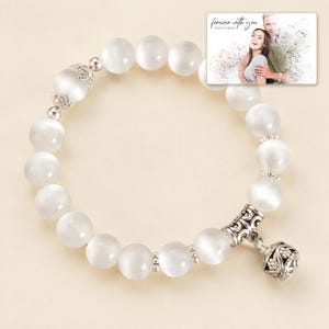May include: A white beaded bracelet featuring smooth, round, semi-translucent beads with a cat's eye effect. The bracelet includes silver-toned metal spacers and a decorative, filigree-style silver charm. A small rectangular photo card shows a couple.