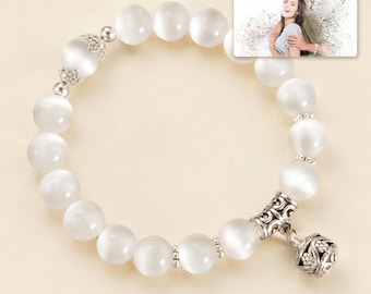 May include: A white beaded bracelet featuring smooth, round, semi-translucent beads with a cat's eye effect. The bracelet includes silver-toned metal spacers and a decorative, filigree-style silver charm. A small rectangular photo card shows a couple.