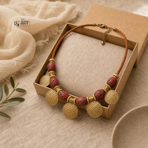 May include: A necklace featuring a brown cord, red porous lava beads, and round, textured brass-toned metal discs. The piece includes small brass-toned spacer beads and a metal lobster clasp closure. The logo GIFT ART DREAM is visible.