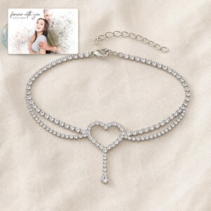 May include: A silver-toned tennis anklet featuring a central heart-shaped charm with a dangling crystal accent. The chain is composed of numerous small, clear, round-cut stones. A small rectangular card displays a photo of a couple with the text "forever with you" and "OLIVIA & ADAM" printed at the top.