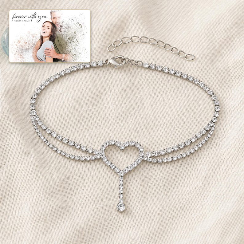 May include: A silver-toned tennis anklet featuring a central heart-shaped charm with a dangling crystal accent. The chain is composed of numerous small, clear, round-cut stones. A small rectangular card displays a photo of a couple with the text "forever with you" and "OLIVIA & ADAM" printed at the top.