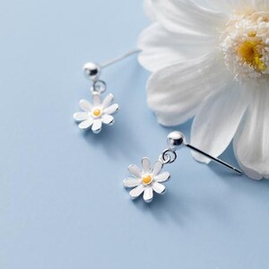 Pair of silver-tone ball stud earrings with dangling white daisy flowers and yellow centers on a pale blue surface.