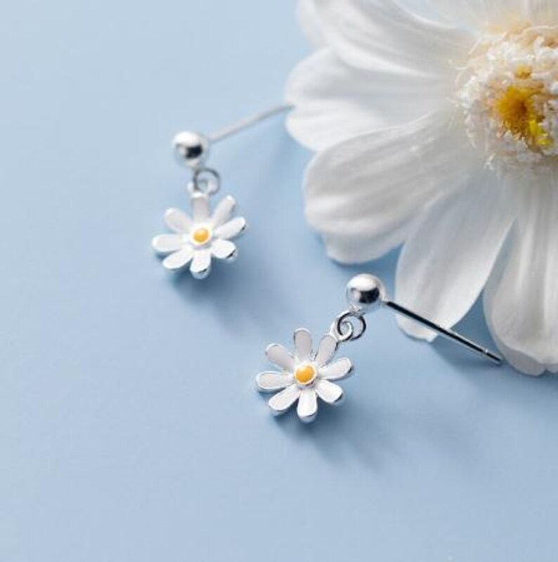 Pair of silver-tone ball stud earrings with dangling white daisy flowers and yellow centers on a pale blue surface.